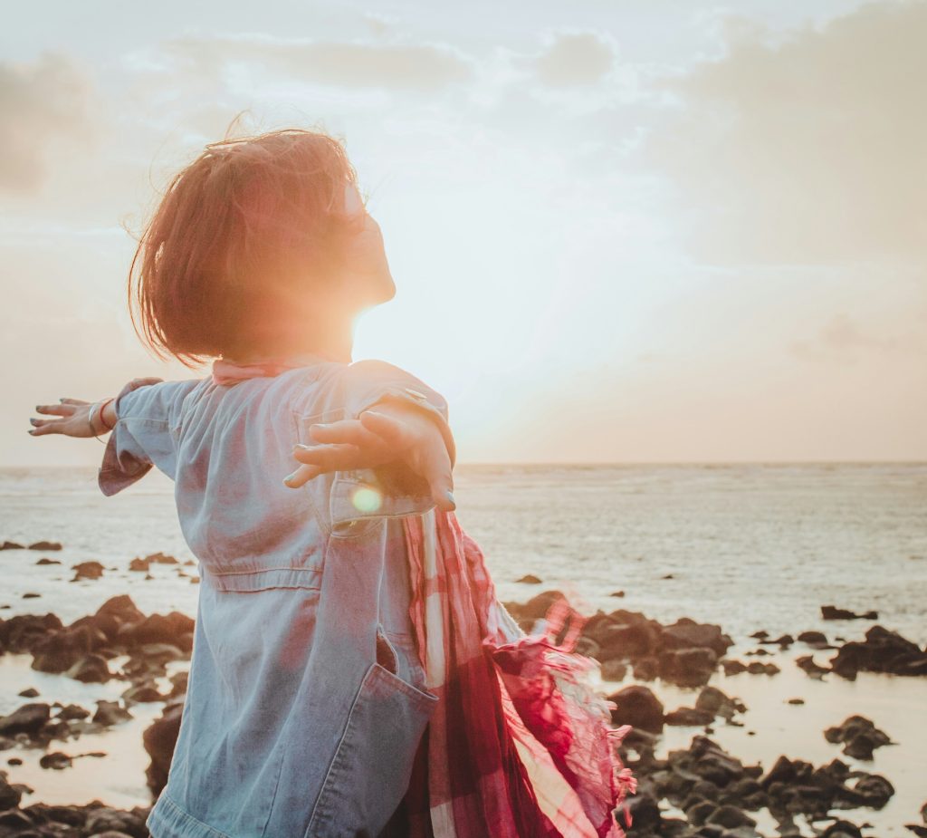 Woman with arms extended to sides in front of water and a sunrise